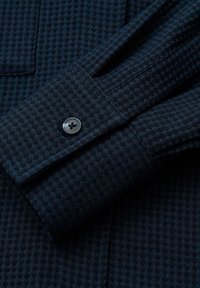 Dark blue shirt cuff featuring a textured, geometric pattern. Includes a round, black button on the sleeve and a pointed cuff design.