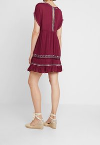 Burgundská šaty s krátkými rukávy, s volánkovým lemem, složitou výšivkou a detaily s klíčovou dírkou na zádech. Spárované s béžovými klíčovými sandály.