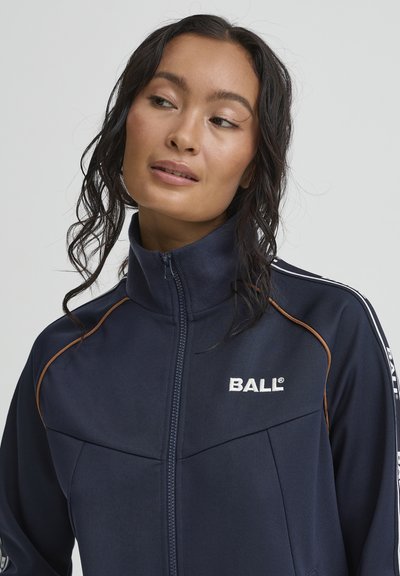 Granatowa kurtka sportowa z wysokim kołnierzem, zamkiem błyskawicznym oraz kontrastującym pomarańczowym przeszyciem. Na piersi znajduje się nazwa marki "BALL" w białym kolorze.