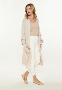 Cardigan lungo fino al ginocchio, aperto davanti, in morbido maglione beige, abbinato a pantaloni bianchi aderenti e ballerine beige chiare. Presenta tasche frontali e polsini a costine.