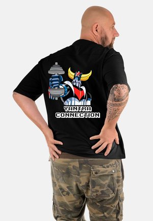Uomo con braccio tatuato che indossa una maglietta nera che mostra la stampa sul retro di un robot dei cartoni animati che tiene un manubrio con la scritta "Yantra Connection", pantaloni mimetici.