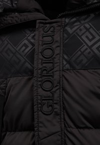Schwarze gepolsterte Jacke mit einem auffälligen "GLORIOUS"-Logo, das auf die Vorderseite gestickt ist, sowie einem strukturierten geometrischen Muster in matter Ausführung.