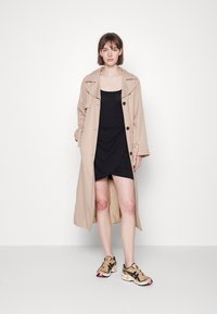Trench coat bege com lapelas largas e botões pretos, usado sobre um vestido preto justo, complementado por ténis bege e pretos.