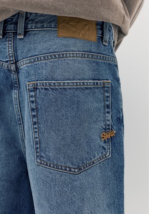 Sininen farkkujen takapuoli, jossa suuri takatasku, ruskea merkkilappu vyötärön yläpuolella ja pieni ruskea logo taskun reunassa.