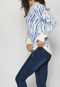 White sweater with blue striped pattern, textured fabric, and contrasting soft cuffs, paired with dark blue jeans featuring decorative stitching.