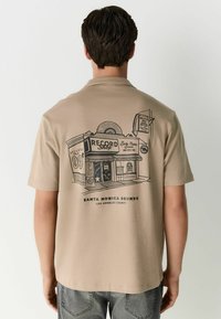Beige short-sleeve shirt featuring a black printed design of a retro record shop on the back with text and graphics. Soft fabric texture.