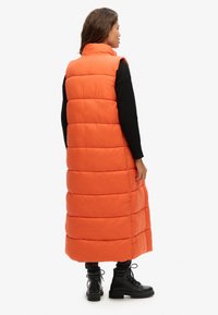 Lange, pluche vest in fel oranje, met horizontaal gewatteerde secties, hoge kraag en een vloer lengte ontwerp. Draagt boven zwarte kleding.