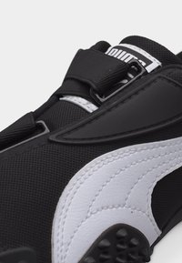 Κοντινή λήψη λευκού και μαύρου αθλητικού παπουτσιού Puma που παρουσιάζει υφασμάτινη υφή, χοντρή λευκή λεπτομέρεια με το λογότυπο και ρυθμιζόμενο λουράκι με αγκράφα.