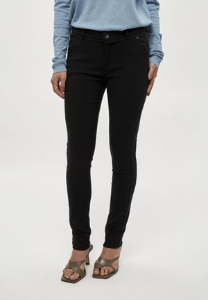 Femme portant un pantalon noir slim, un pull bleu clair et des sandales à talons ouverts, debout devant un fond uni.