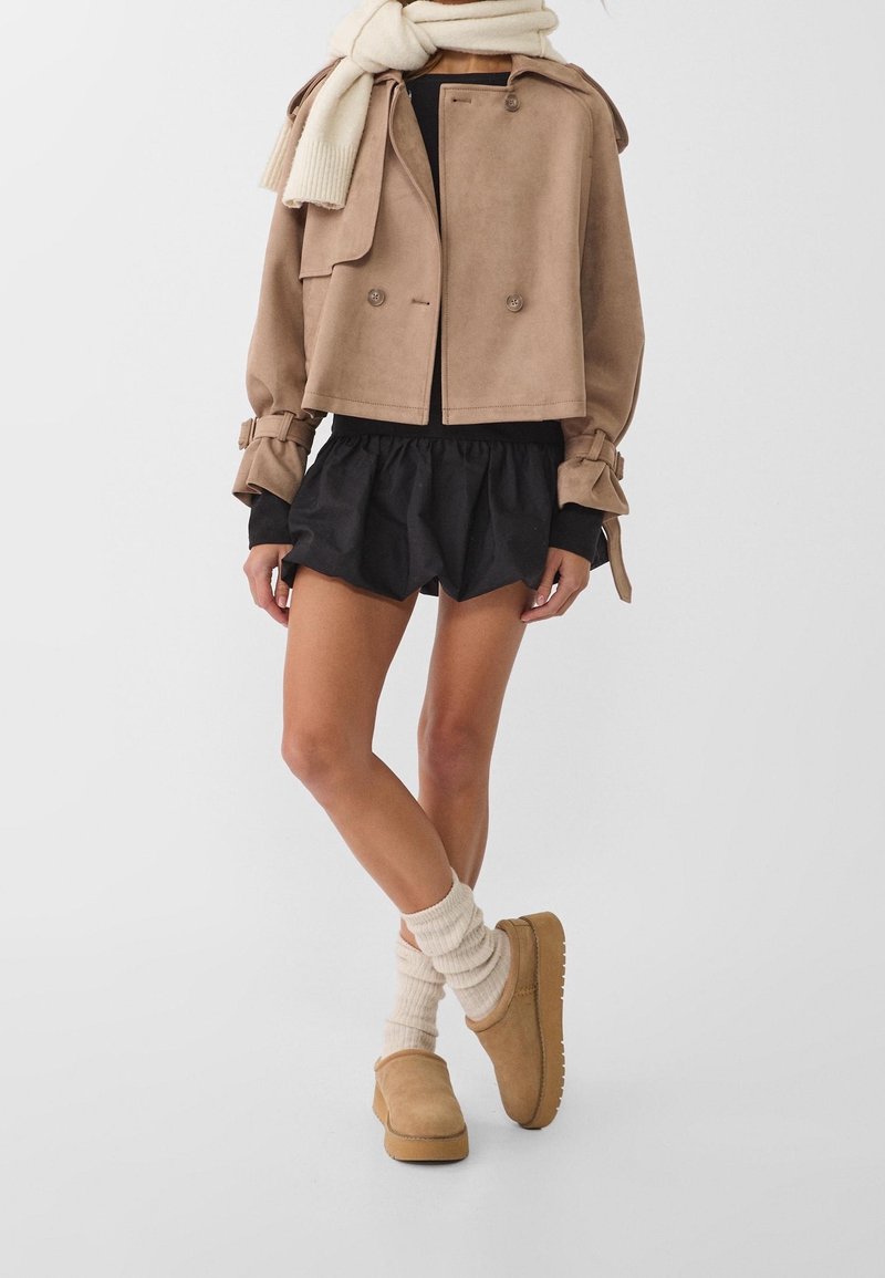 Veste en daim beige coupe courte avec des boutons doubles, shorts noirs à volants, chaussures beige à enfiler et chaussettes côtelées crème jusqu'aux genoux.