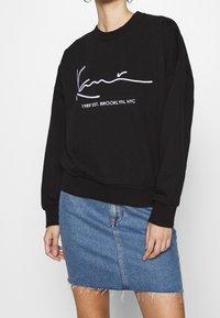 Sudadera negra con cuello redondo y mangas largas. Presenta un texto en blanco con firma y el año impresos en la parte frontal, combinado con una falda de mezclilla.