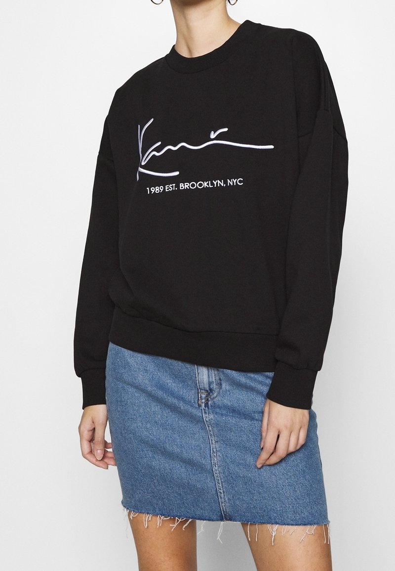 Sudadera negra con cuello redondo y mangas largas. Presenta un texto en blanco con firma y el año impresos en la parte frontal, combinado con una falda de mezclilla.