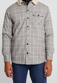 Grå rutiga skjortan med en fuskpälskrage, knappframsida och ett enskilt bröstfack. Tillverkad av tyg med en slät textur och strukturerad design.