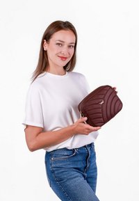Hnědá prošívaná kabelka přes rameno se zipem, zdobená vlnitým vzorem a nenápadným logem. Držena ženou v bílé košili a modrých džínách.