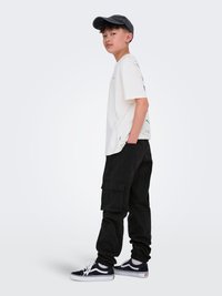 Najstnik z črnim kapom, belim grafičnim majico, črnimi cargo hlačami in črnimi športnimi copati, stoji v sproščenem položaju ob enotni ozadju.