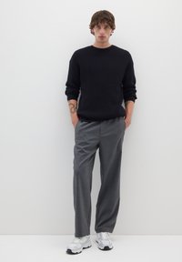 Schwarzer gerippter Pullover, graue, plissierte Hose, weiße Sneakers. Glatte Texturen, lockere Passform und schlichtes Design vor einem einfarbigen Hintergrund.