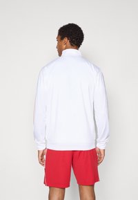 Weiße Sportjacke mit hohem Kragen, langen Ärmeln und roten Akzenten. Kombiniert mit roten Sportshorts. Glatte Textur, lässige Passform.