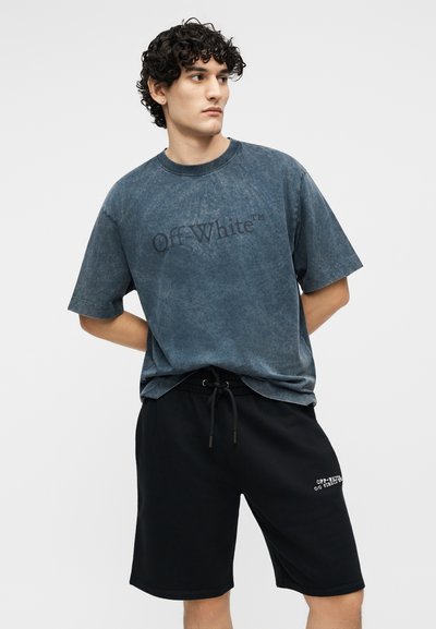 Jeune homme aux cheveux bouclés portant un t-shirt bleu délavé Off-White™ et un short noir à cordon avec du texte blanc sur la jambe gauche.