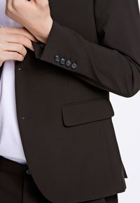 Zwarte blazer met een strak ontwerp, stoftextuur, twee externe zakken en vijf zwarte knopjes op de manchetten. Witte blouse zichtbaar eronder.