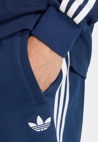 Tmavomodré tepláky s bielymi bočnými pruhmi a malým logom trefoil. Majú vrecko na bedre. Vyrobené z mäkkej bavlnenej látky.