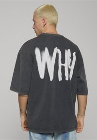 Oversized donkergrijs T-shirt met een losse pasvorm. Witte spuitverftekst "WHY" op de achterkant. Korte mouwen en zachte stoftextuur.