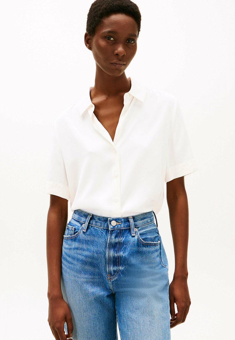 Camicia bianca a maniche corte con bottoni e vestibilità rilassata, abbinata a jeans blu a vita alta, leggermente sbiaditi. Tessuto dalla consistenza morbida.