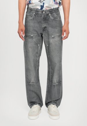 Personne portant un jean noir délavé à jambes droites avec des coutures inclinées distinctives aux genoux et des baskets blanches, debout devant un fond neutre.