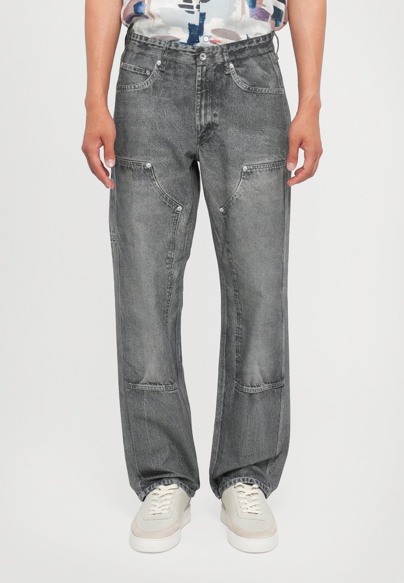 Personne portant un jean noir délavé à jambes droites avec des coutures inclinées distinctives aux genoux et des baskets blanches, debout devant un fond neutre.