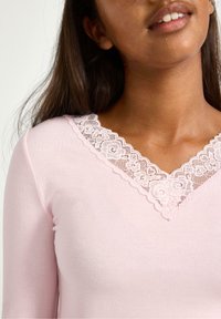 Lichtroze langmouwtop met een V-hals, versierd met delicate witte bloemenkant. Zachte textuur, aansluitend ontwerp.