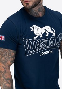 Granatowa bawełniana koszulka z białym logo lwa i napisem "LONSDALE LONDON". Posiada małą naszywkę z flagą Unii na lewym rękawie.