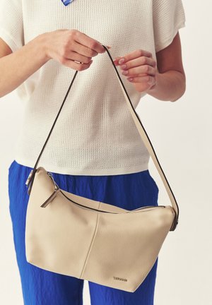 Isik, kes kannab valget tuba ja siniseid pükse, hoiab käes beežist nahast õlakotti, millel on tõmblukk ja tume rihm.