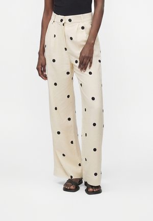 Personne noire portant un pantalon beige à taille haute et jambes larges avec des pois noirs et des sandales noires à bout ouvert, debout sur un fond blanc.