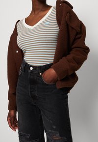 Braune Fleecejacke über einem gestreiften V-Ausschnitt-Shirt in Blau, Braun und Creme, kombiniert mit schwarzen High-Waist-Jeans mit Destroyed-Details.