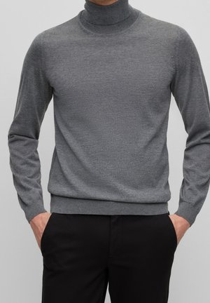 Personne portant un pull gris à col roulé à manches longues et un pantalon noir, mains dans les poches, sur un fond uni.