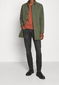 Man i en olivgrön kappa över en rostfärgad tröja, mörkgrå jeans och svarta läderstövlar som står mot en enfärgad bakgrund.