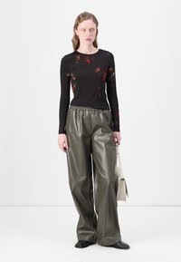Svart langermet topp med blomstertrykk, kombinert med vide grønne bukser i imitert skinn. Modellen holder en kremfarget veske og har på seg svarte sko.