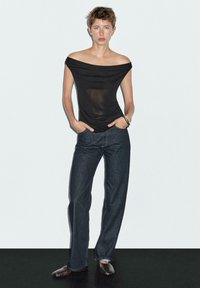 Svart topp med avskuren axel och draperad design, parat med raka mörka jeans och svarta spetsiga skor. Slät textur och figurnära passform.