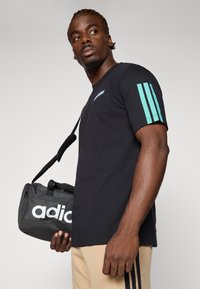 Čierne bavlnené tričko s krátkym rukávom s tyrkysovými trojprúžkami na ľavom rukáve. V kombinácii s čiernou taškou s bielym logom "adidas".