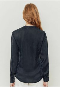 Camicia nera a maniche lunghe con texture setosa, orlo curvo, polsini abbottonati e un sottile dettaglio di cucitura sul retro. Senza marchio visibile.