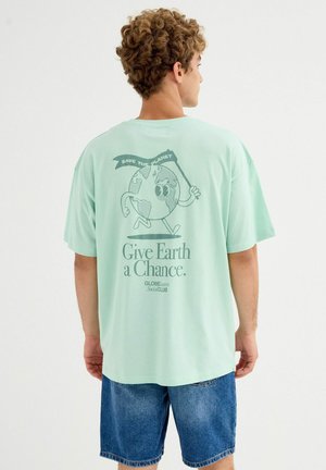 Persona con cabello rizado que lleva una camiseta verde menta mostrando una Tierra corriendo que sostiene una bandera con el texto "Salva el Planeta" y el mensaje "Dale una oportunidad a la Tierra".