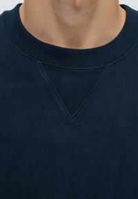 Gros plan d'une personne portant un sweat-shirt bleu foncé à col rond avec un détail côtelé triangulaire sous l'encolure.