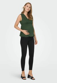 Blouse de maternité vert foncé avec un décolleté en V et une taille froncée, associée à un jean skinny noir avec des détails usés et des fermetures éclair aux chevilles.
