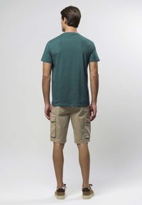 Majica kraćih rukava u tirkiznoj boji uparena s bež cargo kratkim hlačama koje imaju dva bočna džepa. Model nosi smeđe cipele od brušene kože. Pozadina je jednostavna.