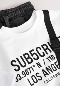 Hvid bomuldssweatshirt med sort tekst, med rund hals. Kombineret med mørkegrå jerseybukser med elastisk talje og snøre.