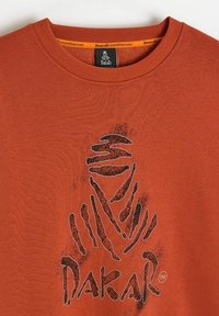 Koszulka w kolorze rdzy z stylizowanym logo Dakar, przedstawiającym owiniętą postać w czarno-białe linie, umieszczoną na środku poniżej kołnierza.