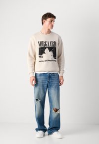 Young man wearing a beige sweatshirt with black print, distressed blue jeans with knee rips, and white sneakers standing against a plain background.