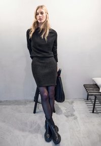 Musta neulepaita, joka on yhdistetty tummanharmaaseen tiukkaan hameeseen, läpinäkyviin mustiin sukkahousuihin ja paksuihin mustiin nilkkasaappaisiin. Yksinkertainen tausta, jossa on puinen jakkara.