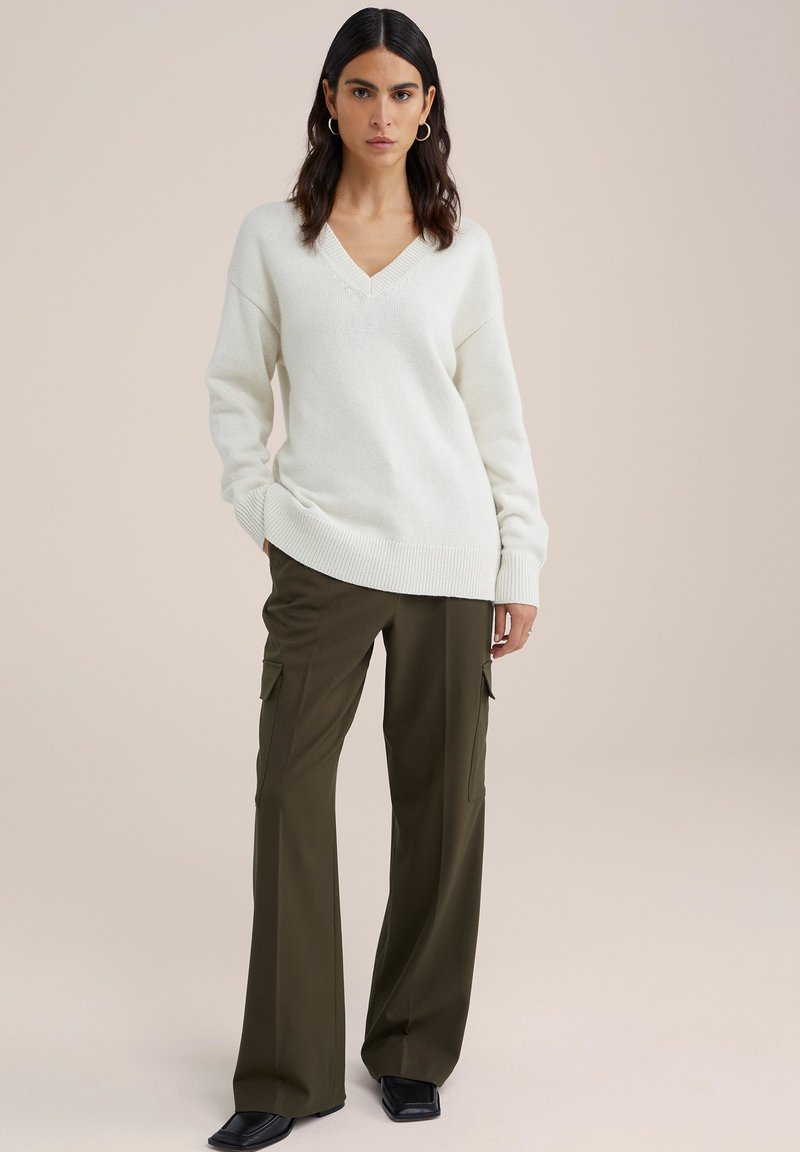 Hellbeige gestrickter Pullover mit V-Ausschnitt und oversized Passform, kombiniert mit olivgrünen Cargo-Hosen mit seitlichen Taschen und lockerer Silhouette.