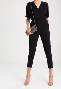 Svart jumpsuit med korta ärmar och V-ringning, med en figurnära midja och avsmalnande cropped ben. Matchad med en liten grå clutch.