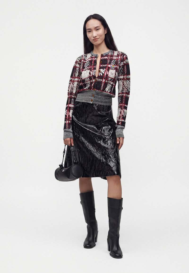 Femme portant un cardigan court à carreaux rouge et noir, une jupe noire brillante au genou, des bottines noires à talons, et tenant un sac à main noir.